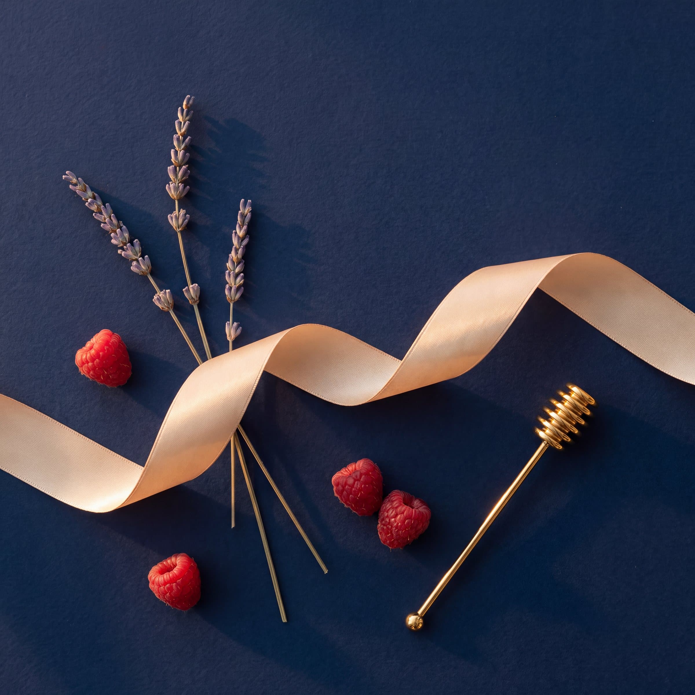 Ruban champagne, brins de lavande, framboises et cuillère à miel dorée sur papier bleu nuit